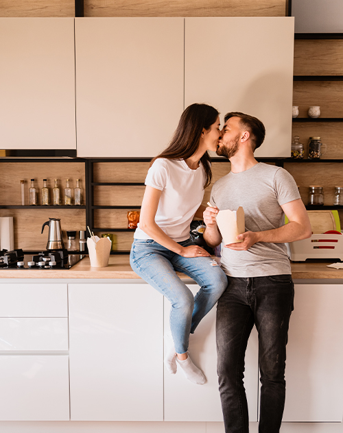 Couple souriant, tenant des tasses de café, dans une cuisine de style classique avec des meubles blancs et une crédence en briques. Illustration d'un aménagement de cuisine par Bricomarché Cosne."