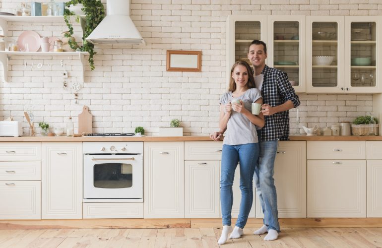 Couple souriant, tenant des tasses de café, dans une cuisine de style classique avec des meubles blancs et une crédence en briques. Illustration d'un aménagement de cuisine par Bricomarché Cosne."